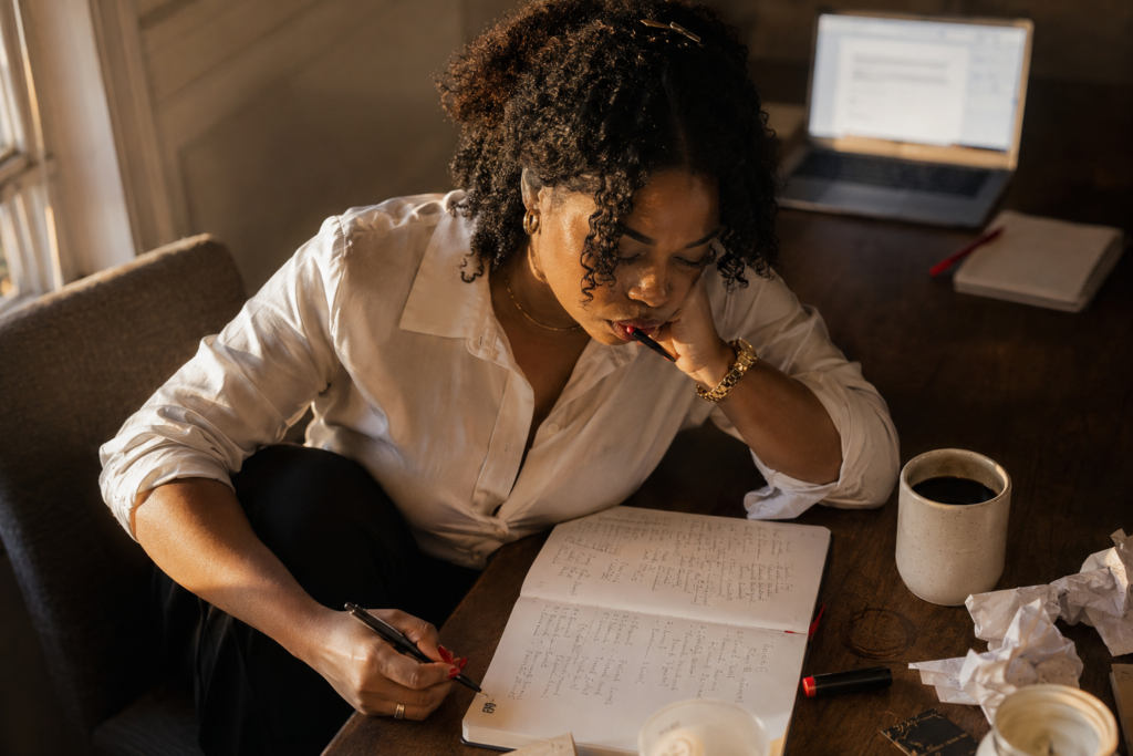 Black woman with natural curly hair partially pinned up with a gold clip, wearing a white button-down shirt with sleeves rolled, leaning over a dark wood table writing intensely in an open journal filled with dense handwritten notes, right hand writing with red nails visible, left hand with a gold chain bracelet holding a pen to her lips in a thinking pause, ceramic coffee mug and crumpled papers beside the journal, open laptop with a document visible in the upper background, warm natural window light from the left illuminating the scene. Her SoftPower Astrology.