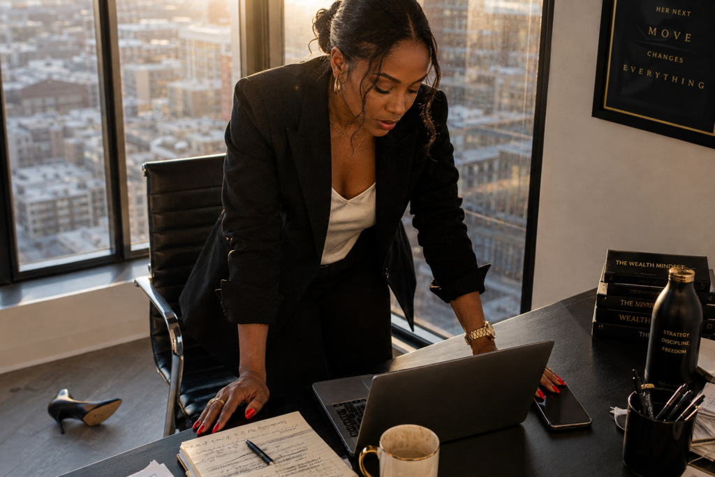 Black woman in a black blazer and white blouse leaning over a dark desk in a high-rise office at golden hour, red nails and gold watch visible as she reviews an open laptop, handwritten journal and gold-handled coffee mug on the desk, stacked books reading The Wealth Mindset and a black water bottle reading Strategy Discipline Freedom to the right, a framed poster on the wall reading Her Next Move Changes Everything, floor-to-ceiling windows with a city skyline behind her, heels kicked off on the floor. Her SoftPower Astrology. Astrology journal ideas for Black women. Birth chart alignment. Dating advice for women 30s and 40s.